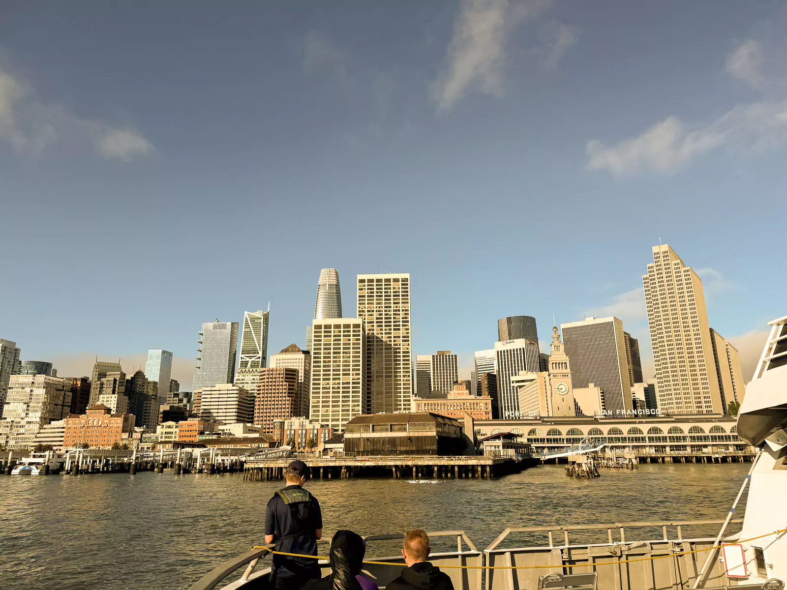 A view of downtown San Francisco’s skyline and the historic Ferry Building seen from a ferry on the water. The bustling waterfront area includes piers and boats under a clear sky.