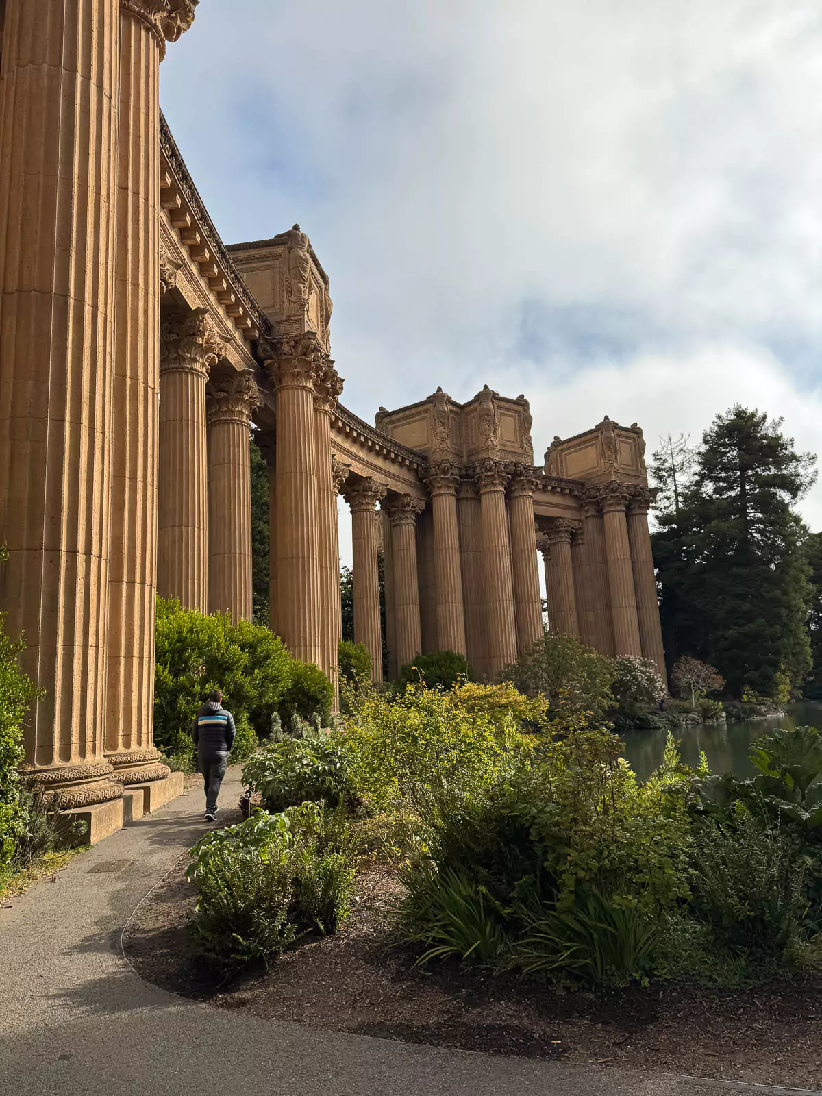 Colonnade at the Palace of Fine Arts in San Francisco, featuring towering Corinthian columns and ornate detailing. The classical architecture is surrounded by greenery and reflected in the tranquil lagoon nearby, creating a peaceful and grand setting.