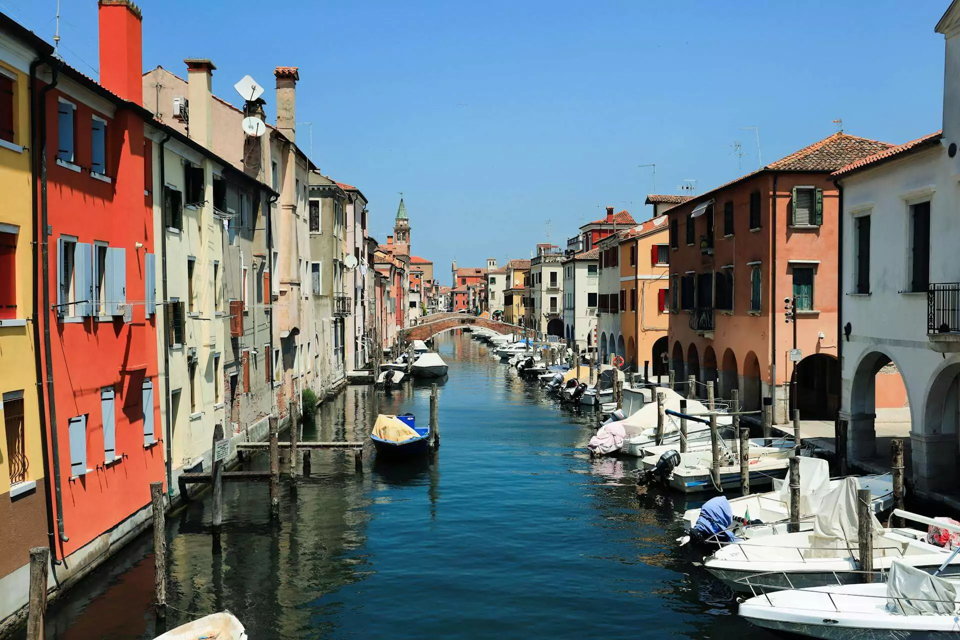 Canal scene in Chioggia, Italy, with colorful historic buildings lining the waterway, small boats docked along the edges, and a stone bridge in the background