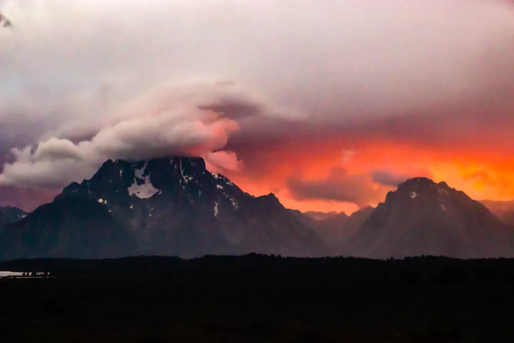 Vibrant bright orange clouds glow above rugged mountain peaks near Jackson Lake Lodge at sunset, casting a warm light over the landscape. The dramatic sky contrasts with the darkening silhouette of the mountains.