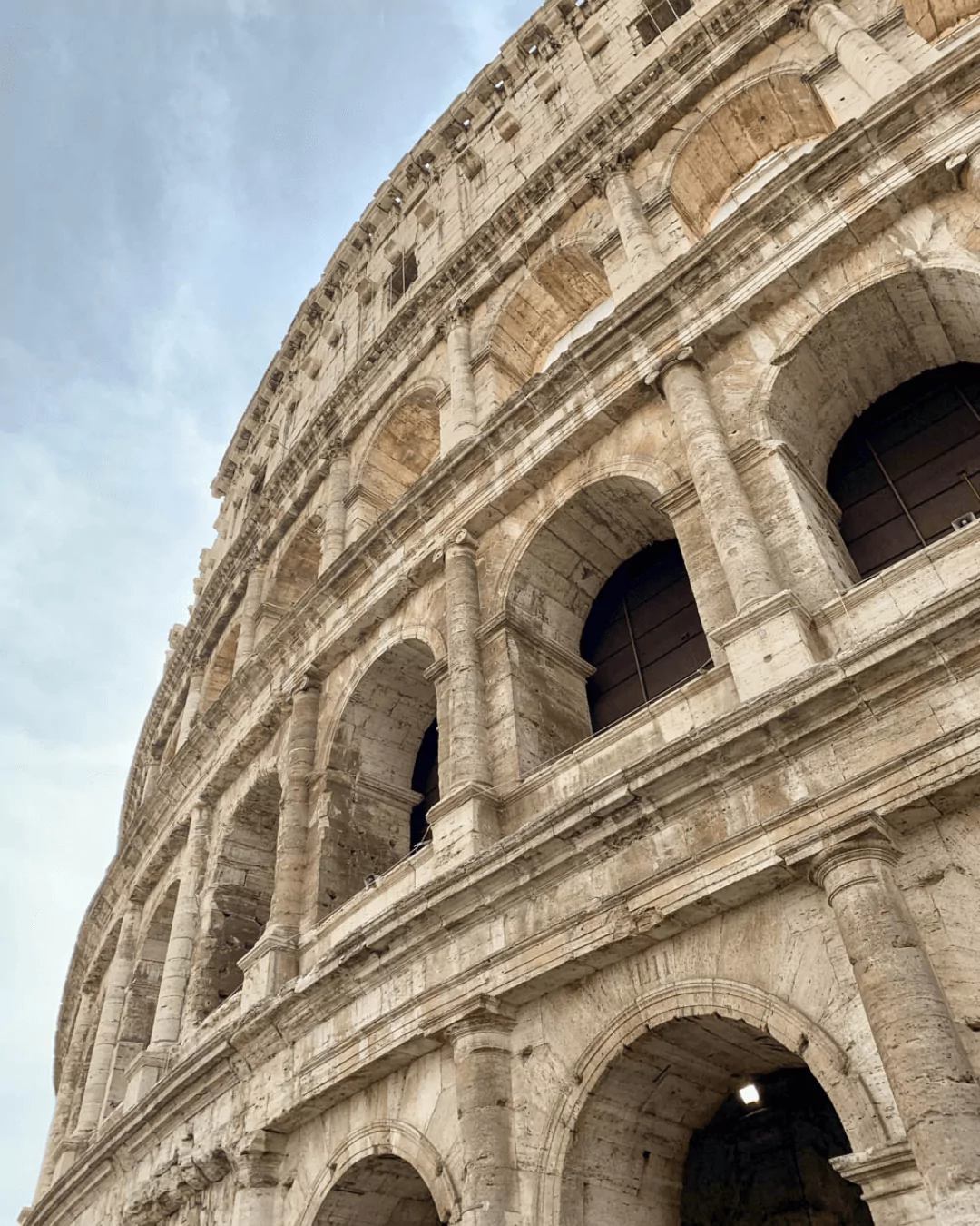 Angled view of the Colosseum's outer walls in Rome, showcasing the towering arches and weathered stonework of the ancient amphitheater. The perspective highlights the scale and grandeur of this iconic Roman landmark against a bright sky.