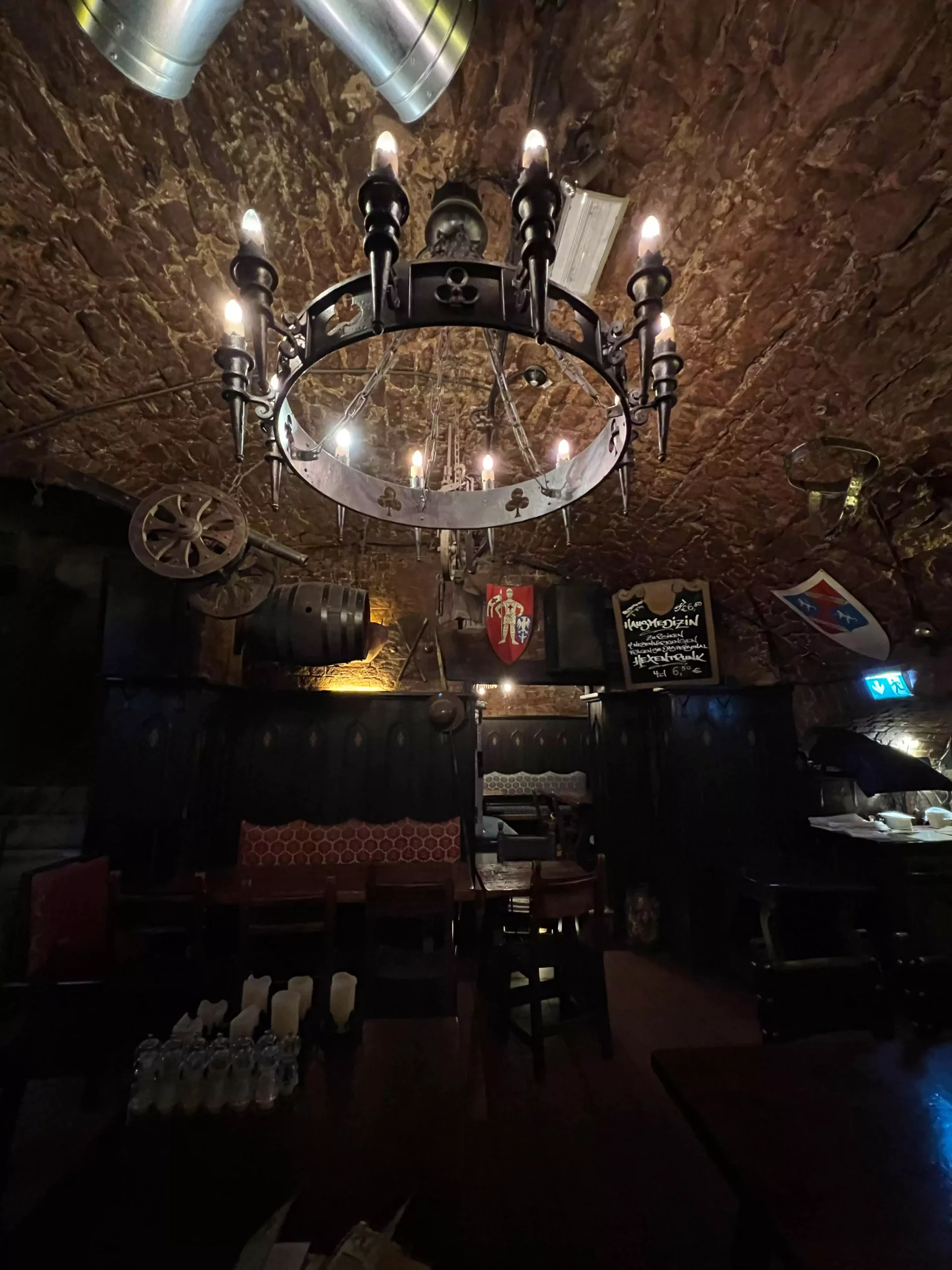 The dark, rustic interior of Im Keller in Nuremberg, Germany, featuring stone walls, wooden beams, and a striking iron chandelier hanging from the ceiling. The warm, moody lighting enhances the historic cellar atmosphere.
