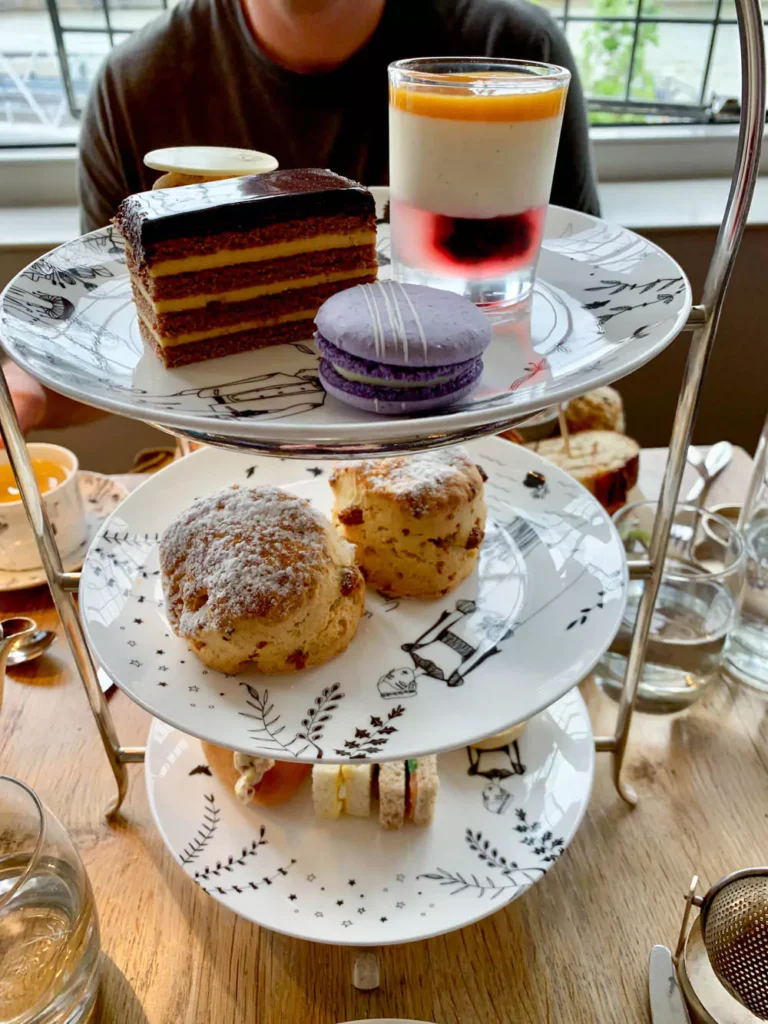 A selection of classic afternoon tea treats including finger sandwiches, scones with clotted cream and jam, and assorted pastries arranged on a tiered stand. A teapot and fine china cups accompany the spread, highlighting a traditional British tea experience.