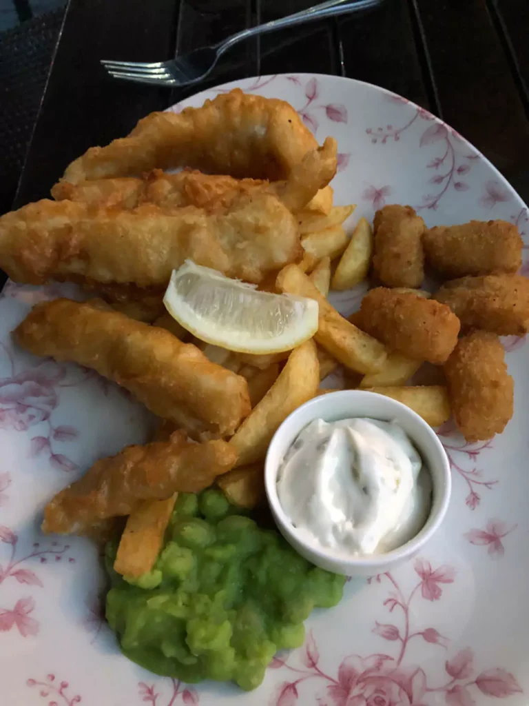 Classic British fish and chips served at a pub in London, featuring golden battered fish, thick-cut fries, and a side of mushy peas. The hearty dish is plated on a white dish, capturing a staple of traditional UK pub fare.