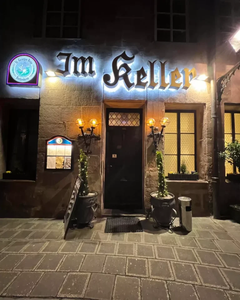 Nighttime view of the exterior entrance to Im Keller in Nuremberg, Germany, with warm light spilling from the doorway onto the cobblestone street. The stone facade and wooden door create an inviting, old-world ambiance.
