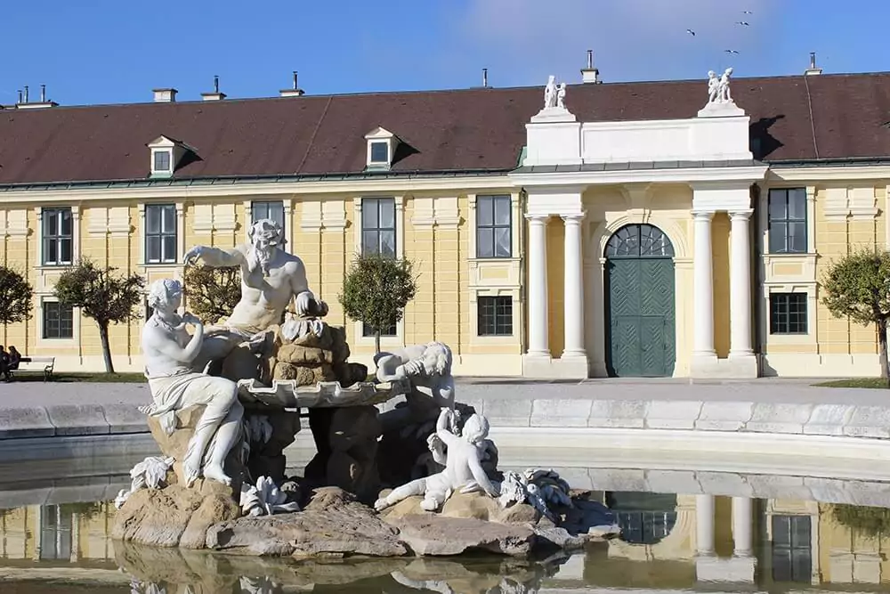 A grand fountain at the entrance to Schönbrunn Palace in Vienna features sculpted figures and flowing water set in a circular stone basin. The baroque design and elegant symmetry welcome visitors with classic imperial flair.