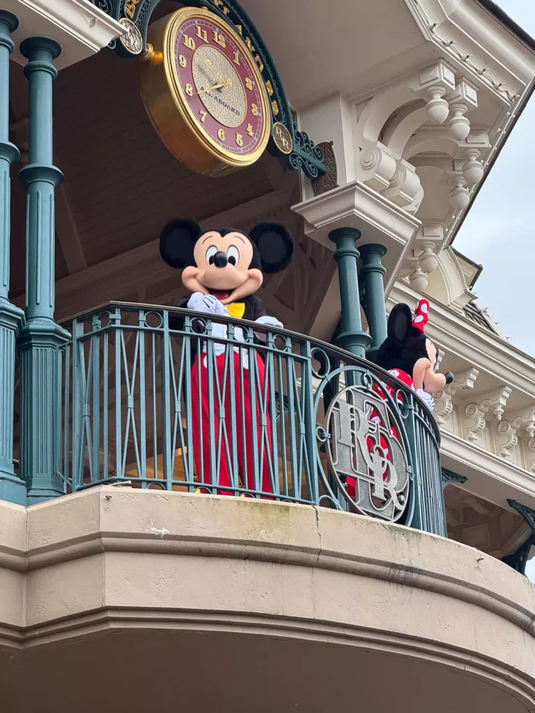 Minnie and Mickey Mouse waving happily to guests, dressed in their classic red, black, and yellow outfits, creating a joyful and welcoming scene at a Disney park.