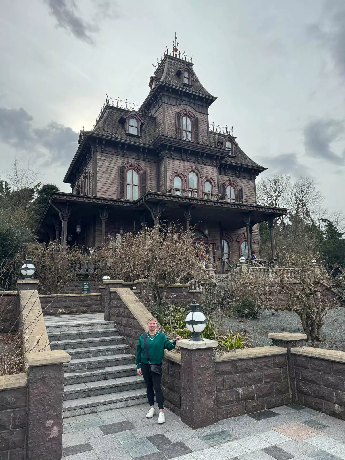 Katie standing in front of Phantom Manor at Disneyland Paris, smiling with the spooky, old Western-style haunted mansion rising behind her under a slightly cloudy sky.