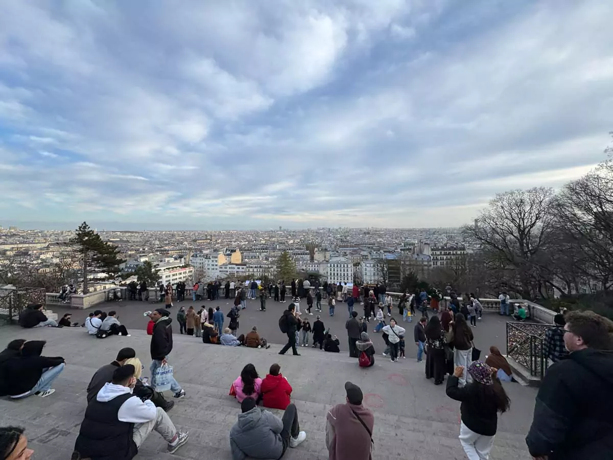 Sweeping view of Paris from the top of Sacré-Cœur, with the city’s rooftops stretching into the distance, iconic landmarks peeking through the skyline, and soft light casting a golden glow