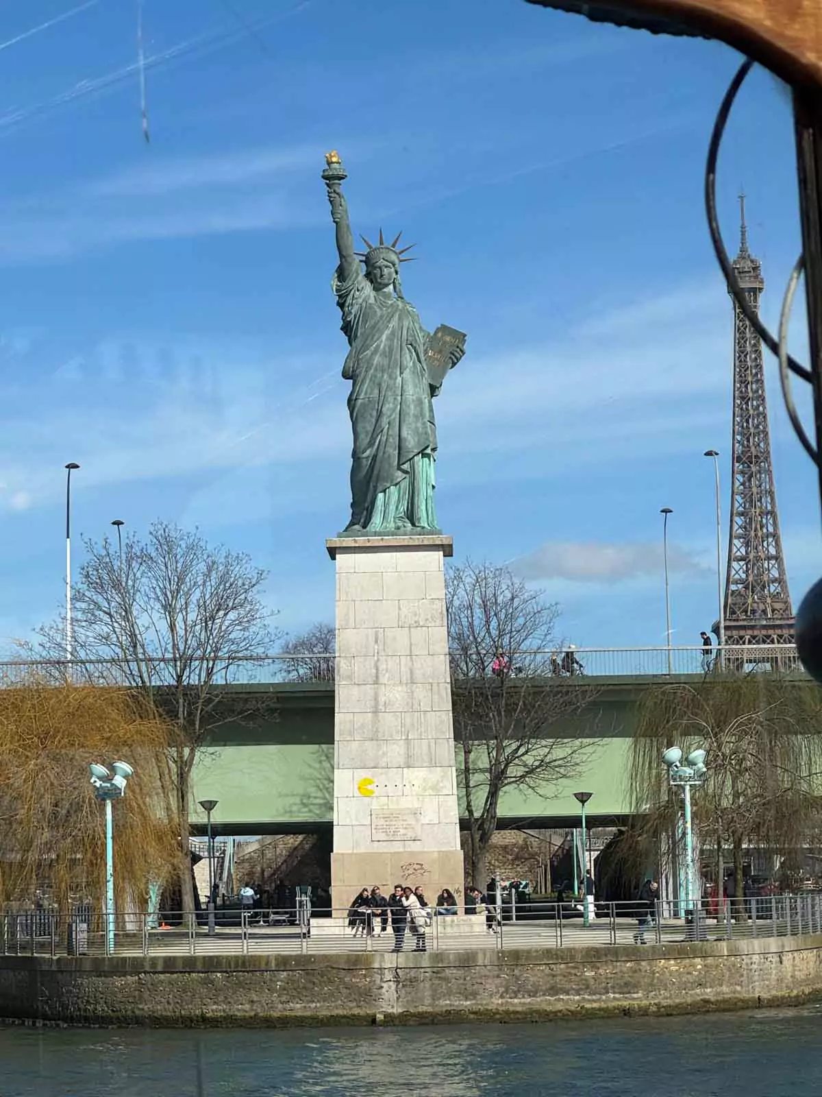View of the replica Statue of Liberty along the Seine River in Paris, seen from the deck of the Calife lunch cruise boat. The iconic monument stands proudly with the Eiffel Tower faintly visible in the distance, blending French and American symbols during a scenic river journey.