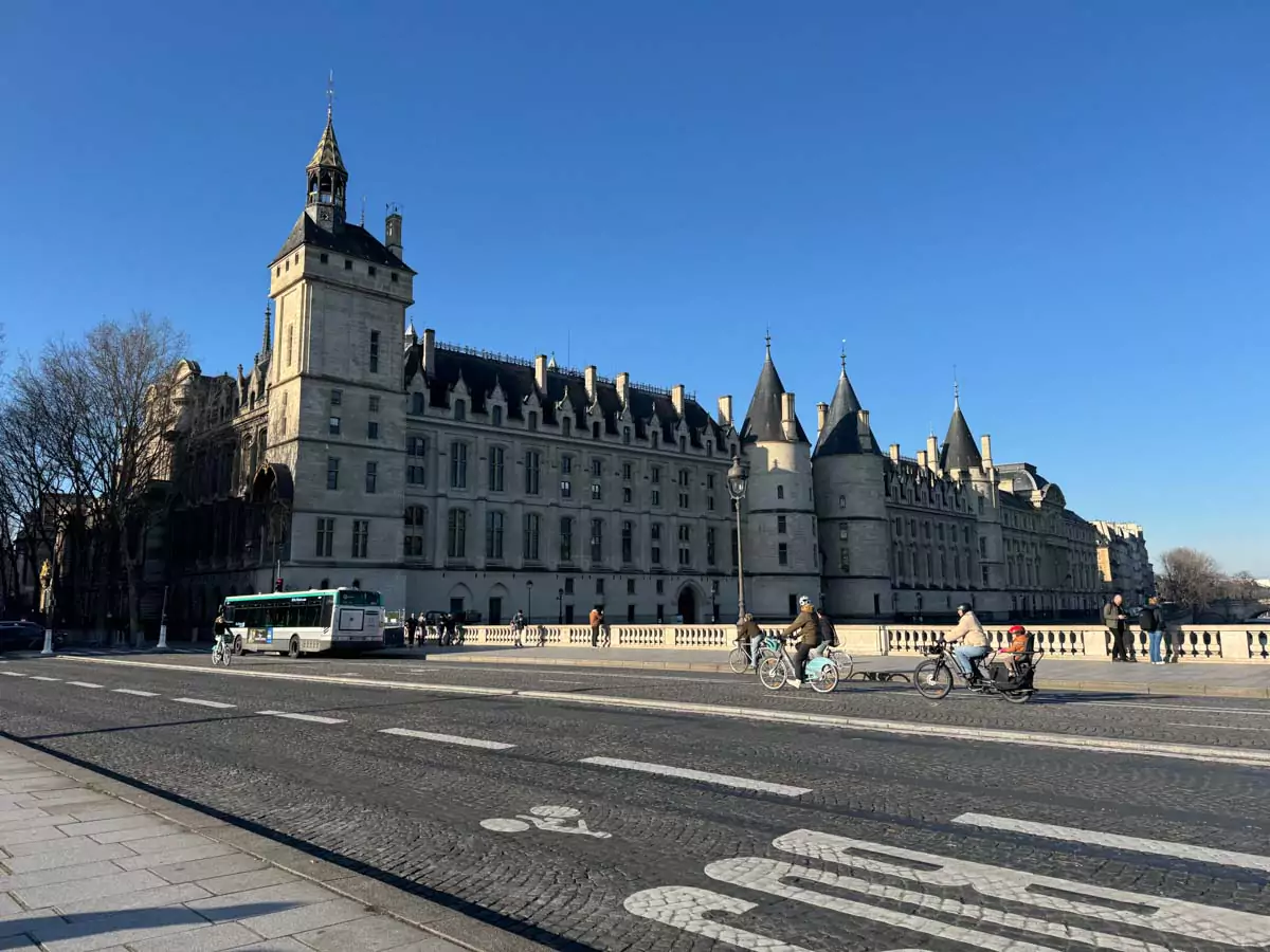Exterior of the Conciergerie in Paris, France, seen from across the Seine River, featuring its medieval stone towers, pointed turrets, and grand historic facade. The former royal palace and prison stands as a striking landmark on the Île de la Cité.