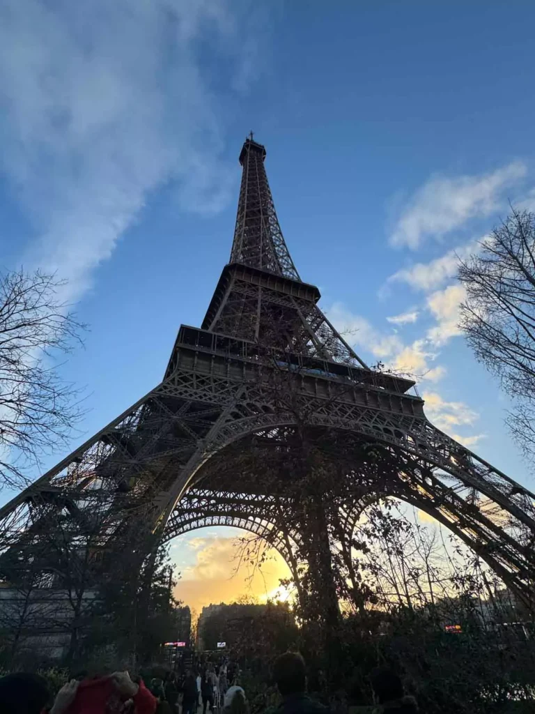 Daytime view of the Eiffel Tower from its base in Paris, France, looking up through the iron lattice structure as sunlight casts shadows on the ground — an awe-inspiring angle