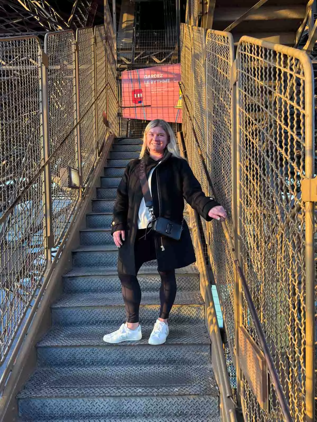 Katie climbing the stairs of the Eiffel Tower in Paris, France, with the tower’s intricate ironwork surrounding her and the city skyline peeking through — a candid and adventurous moment