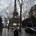 Katie standing on a charming Parisian side street with the Eiffel Tower rising in the background, framed by classic Haussmann buildings and soft daylight — a stylish city moment