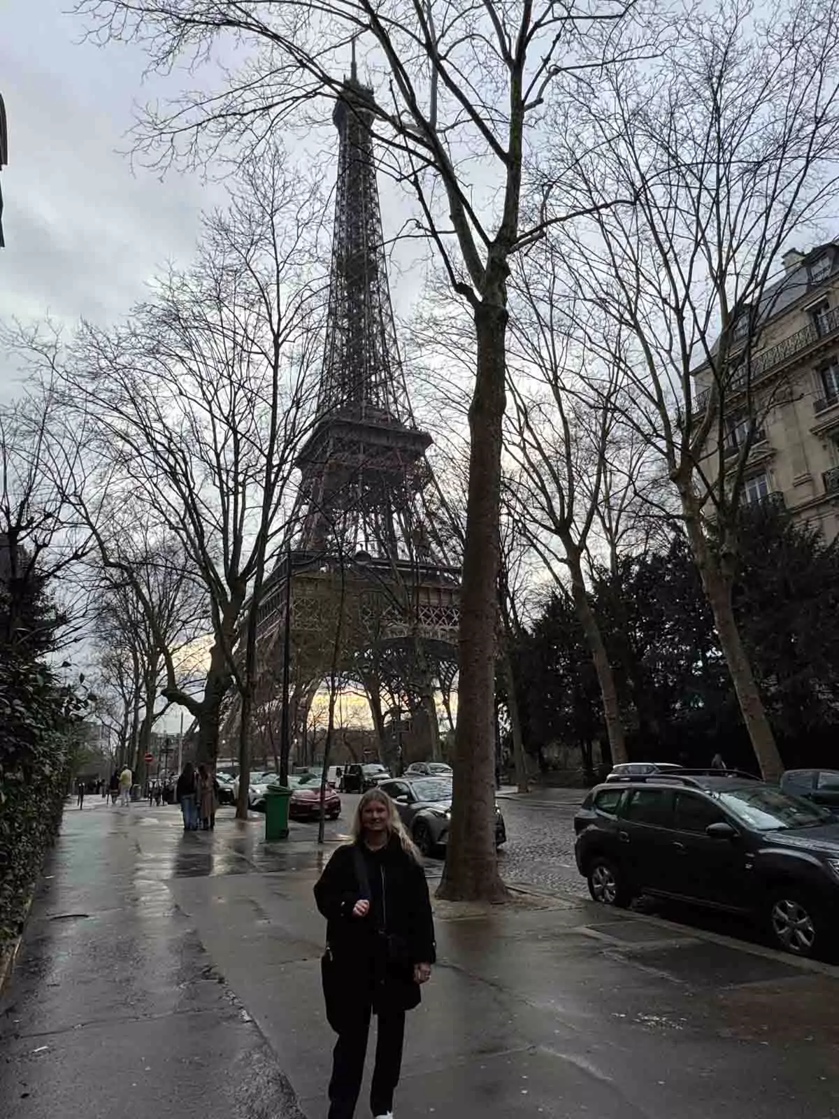 Katie standing on a charming Parisian side street with the Eiffel Tower rising in the background, framed by classic Haussmann buildings and soft daylight — a stylish city moment