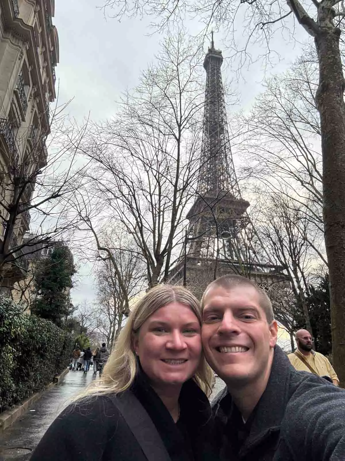Katie and Zack standing on a quiet Parisian side street with the Eiffel Tower in the background, surrounded by classic architecture and soft evening light — a romantic city moment