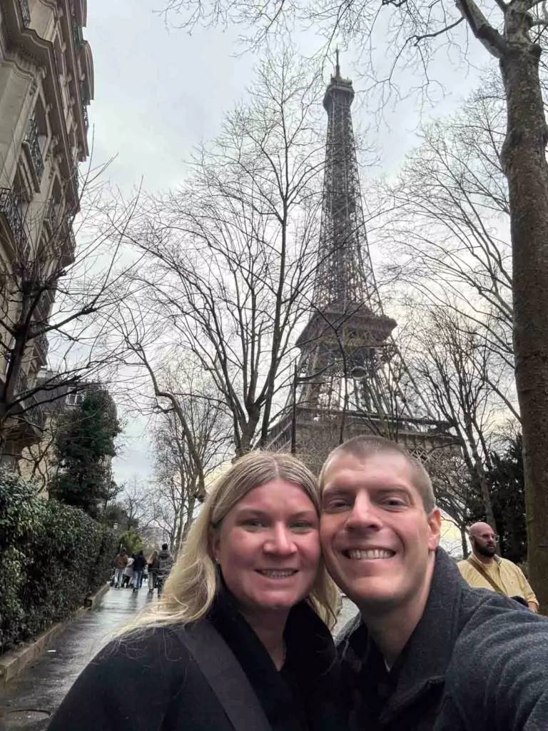 Katie and Zack taking a selfie in front of the Eiffel Tower in Paris, France, smiling with the iconic iron landmark towering behind them and a soft Parisian sky above.