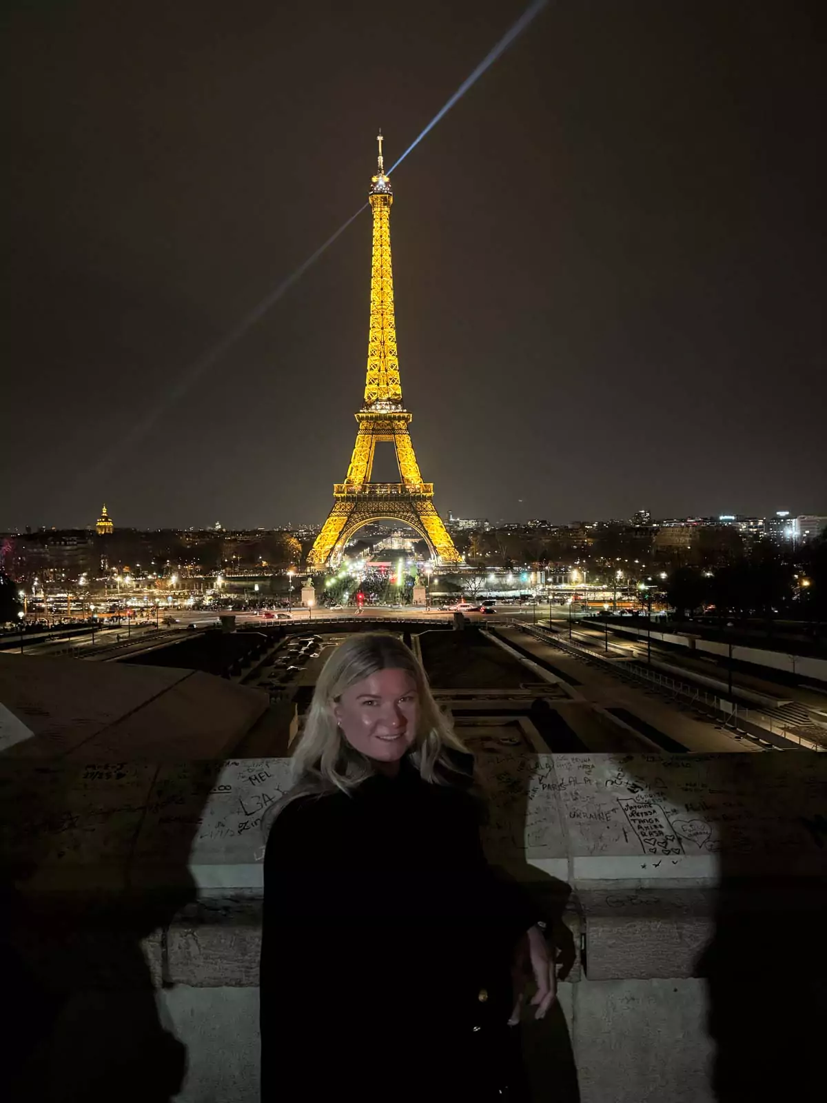 Katie admiring the Eiffel Tower at night from the Trocadéro in Paris, France, with the tower glowing in golden lights and reflections shimmering on the wet plaza — a magical nighttime view