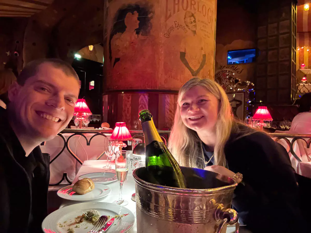 Katie and Zack seated at a dinner table inside the Moulin Rouge in Montmartre, Paris, smiling and dressed for an elegant night out, surrounded by the cabaret’s iconic red ambiance and lively atmosphere.