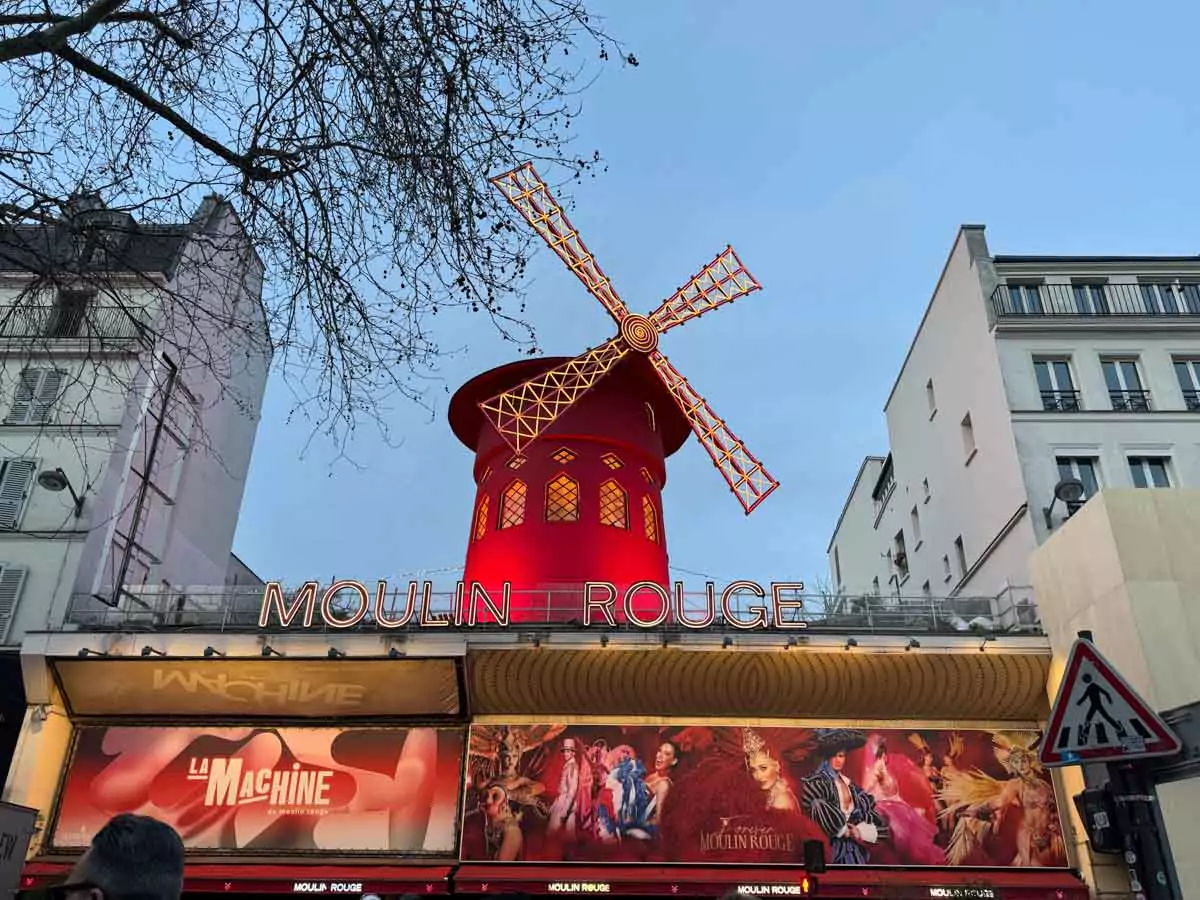 Exterior of the Moulin Rouge in Montmartre, Paris, featuring its iconic red windmill atop the entrance, glowing neon signage, and lively atmosphere at one of the city’s most famous cabarets.