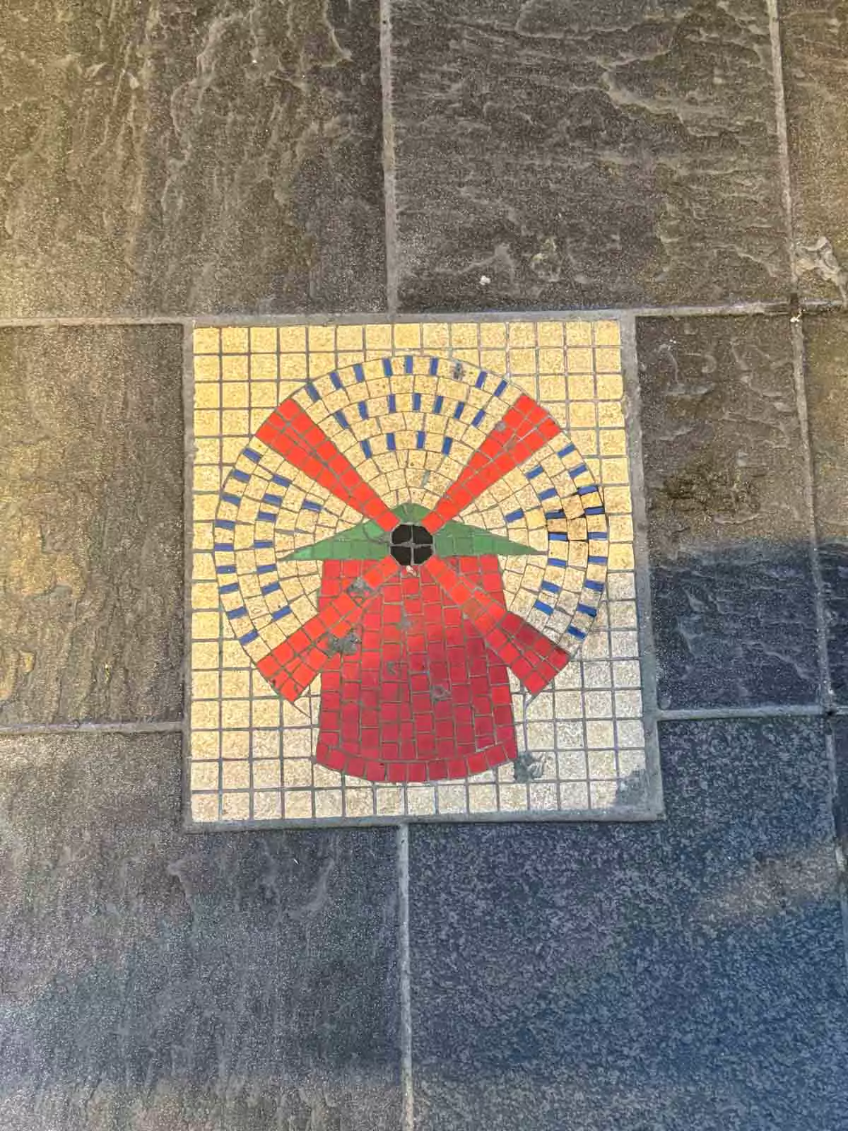 Decorative tile on the sidewalk near the Moulin Rouge in Montmartre, Paris, featuring a stylized red windmill design, paying tribute to the iconic cabaret’s emblem in a subtle street detail.