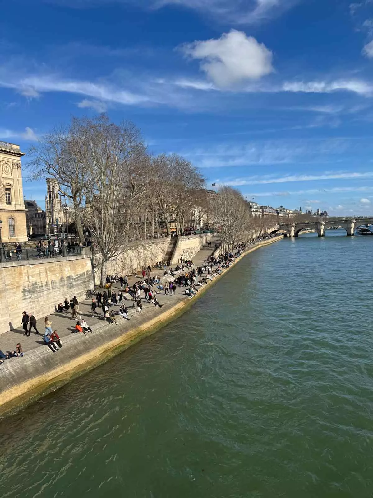 People sitting along the edge of the Seine River in Paris, France, enjoying a relaxed moment with views of the water, passing boats, and classic city architecture — a laid-back afternoon scene