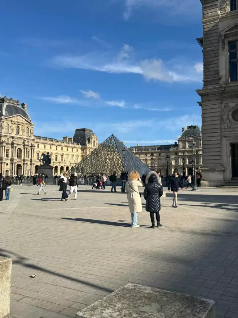 The glass pyramid entrance of the Louvre Museum in Paris, France, with visitors gathered around the modern structure framed by the historic palace wings — a striking blend of old and new