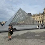 Katie standing near the glass pyramid entrance of the Louvre Museum in Paris, France, with the modern structure reflecting the sky and framed by the classical museum buildings — a stylish arrival