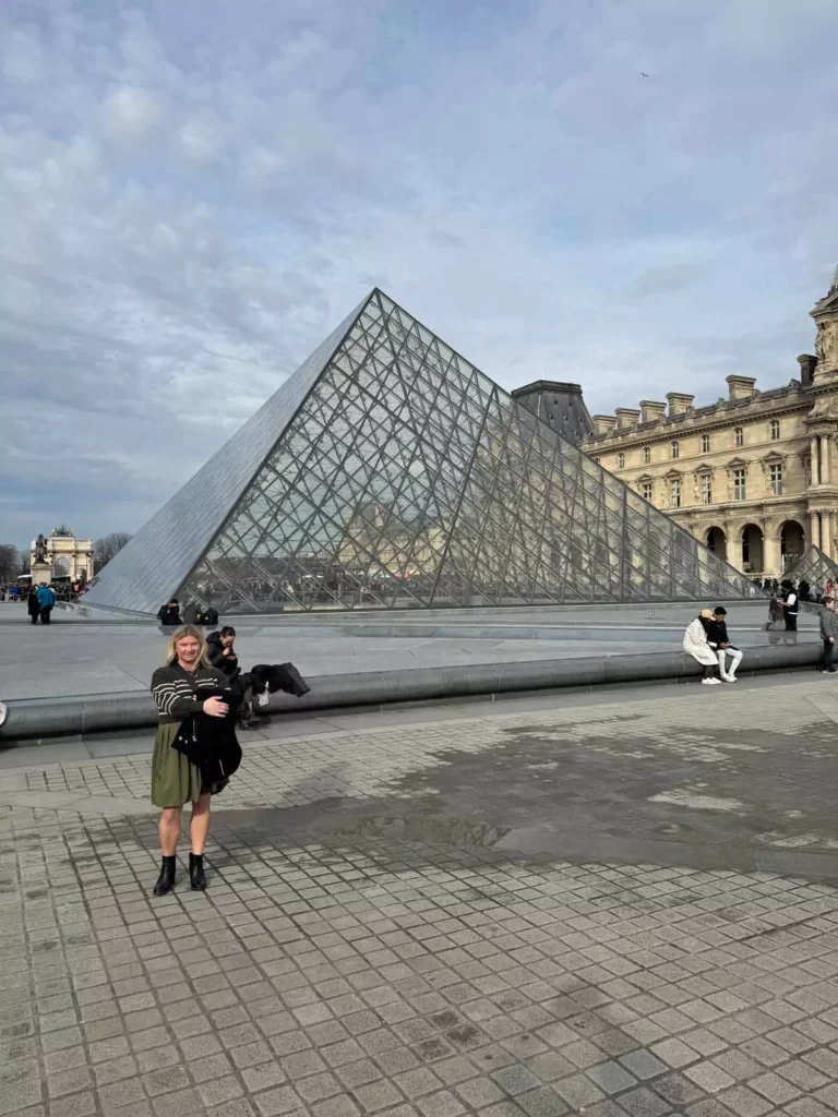 Katie standing near the glass pyramid entrance of the Louvre Museum in Paris, France, with the modern structure reflecting the sky and framed by the classical museum buildings — a stylish arrival