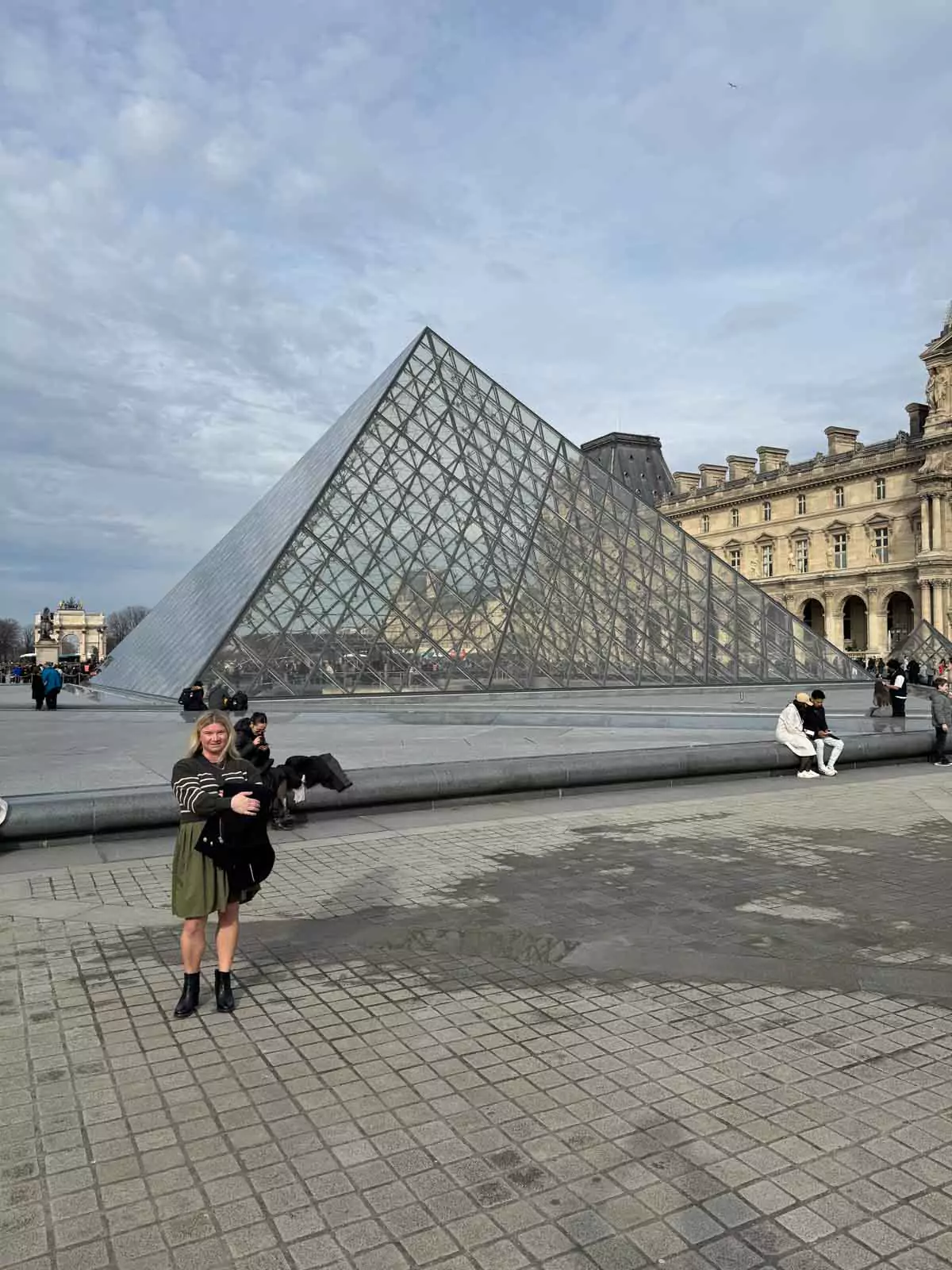 Katie standing near the glass pyramid entrance of the Louvre Museum in Paris, France, with the modern structure reflecting the sky and framed by the classical museum buildings — a stylish arrival