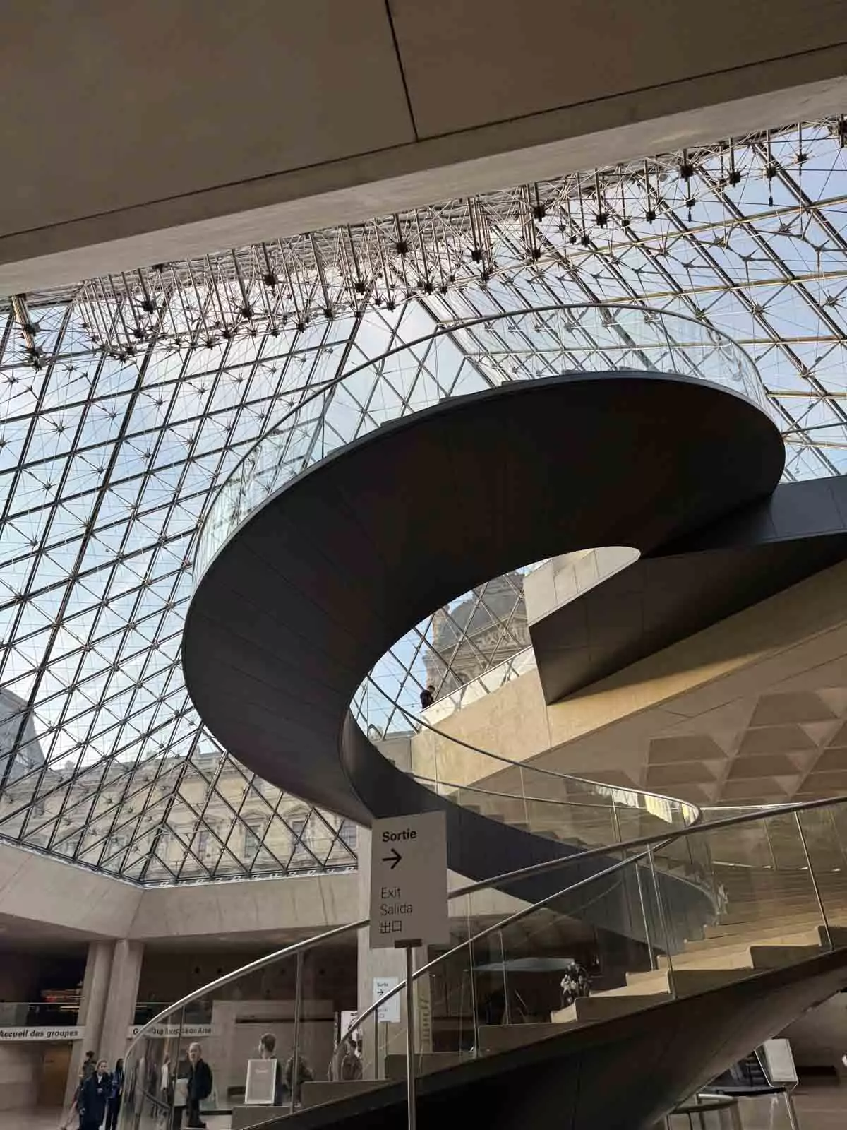 View of the Louvre pyramids from inside the museum, looking up from the spiral staircase with glass panels above and visitors circling below