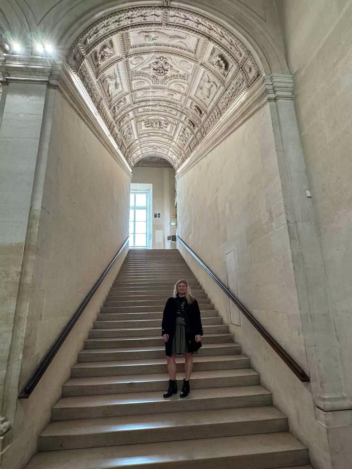 Katie ascending the Henri II staircase at the Louvre Museum in Paris, France, surrounded by sculpted stone walls and a vaulted ceiling with ornate ribbing — a graceful blend of movement and architecture