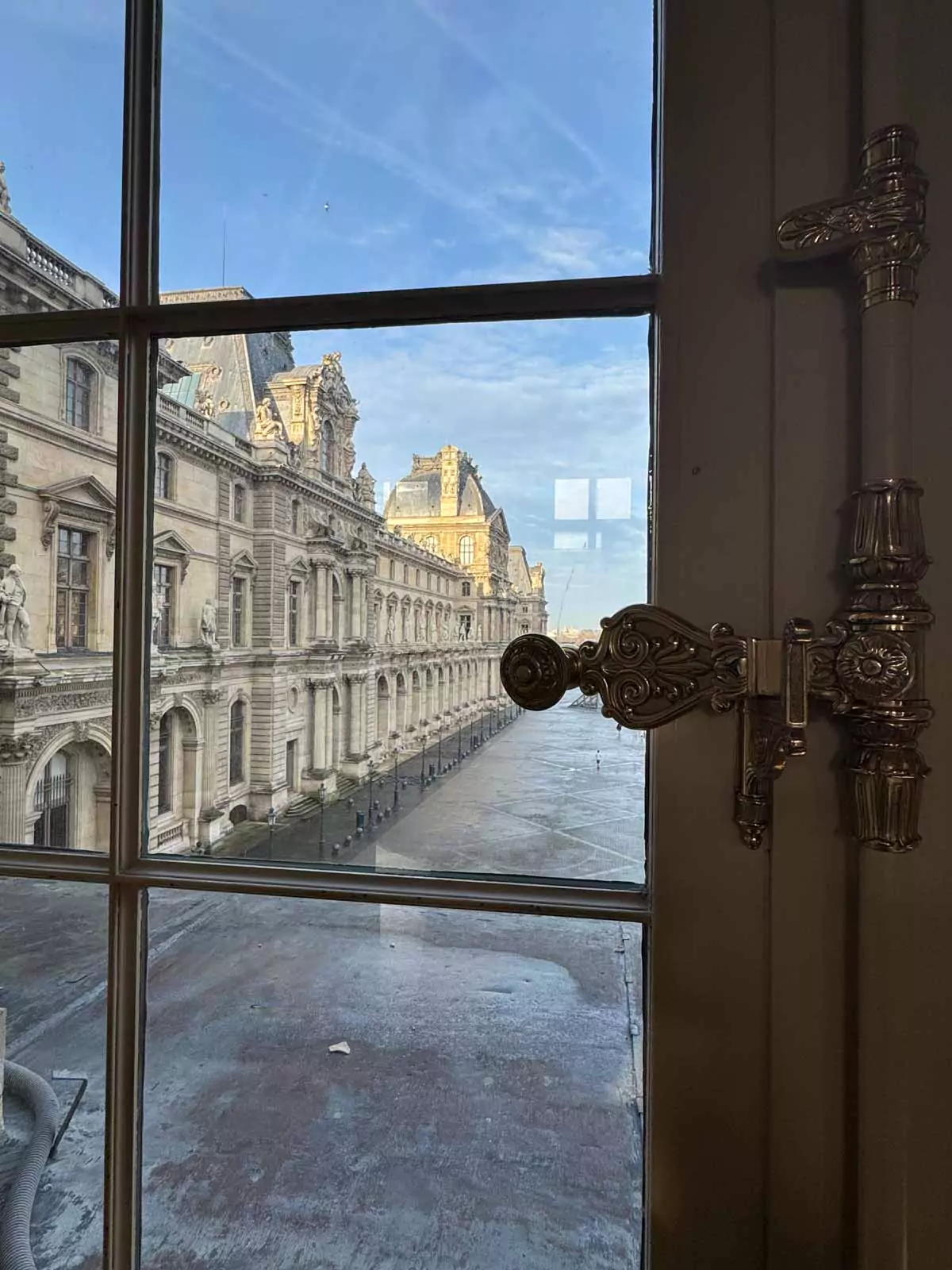 View from inside the Louvre Museum in Paris, France, looking out through a grand window onto the courtyard and glass pyramid, with natural light casting soft shadows on the stone interior