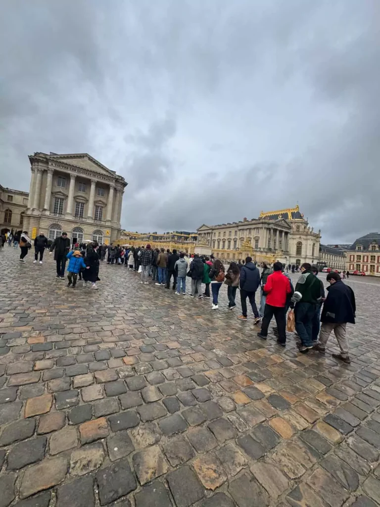 Long line of visitors waiting at the entrance to the Palace of Versailles, stretching past the golden gates under a sunny sky, with tourists prepared for security checks and ticket scanning — a typical arrival scene at versailles