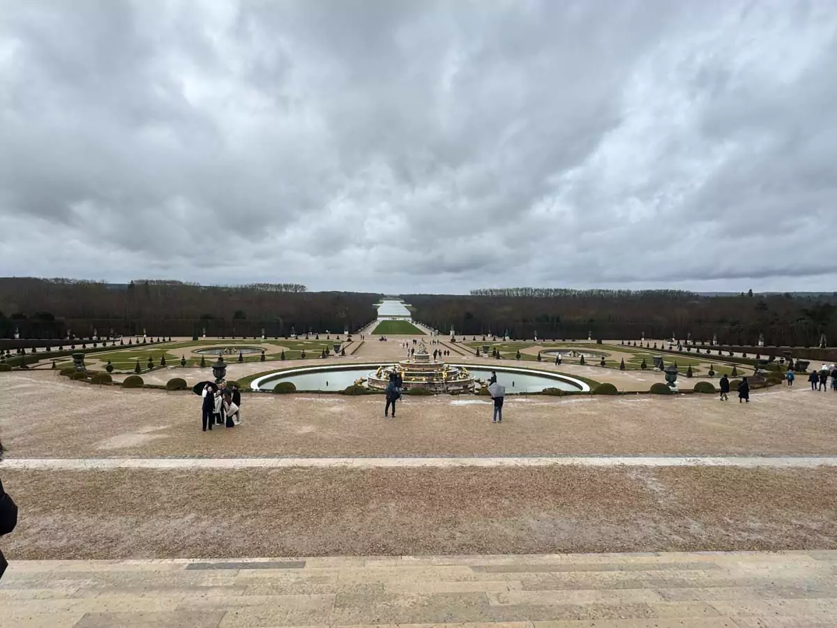 A grand view from the Palace of Versailles across the formal gardens, with symmetrical hedges, fountains, and gravel paths leading the eye straight toward the distant lake