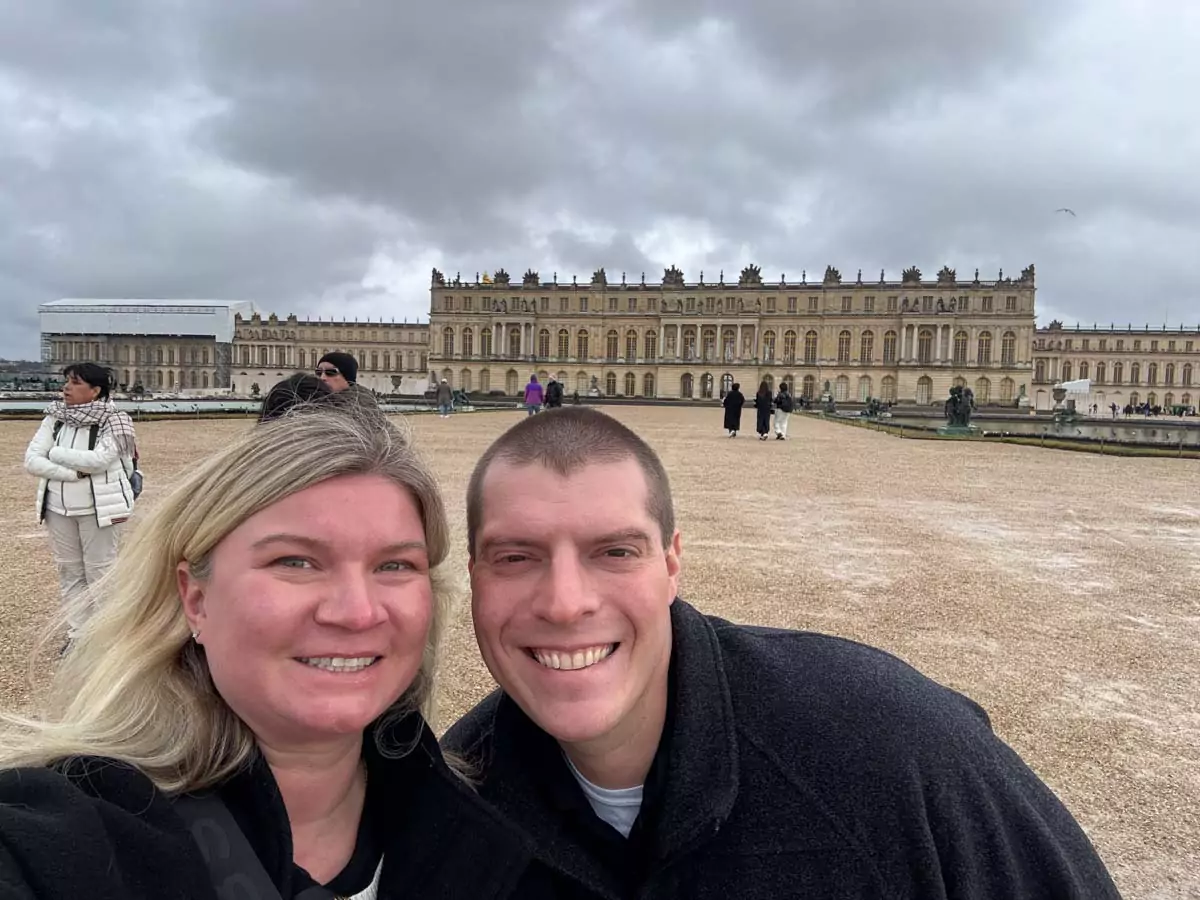 Katie and Zack walking through the Palace of Versailles gardens, with manicured hedges and fountains surrounding them and the grand façade of the palace rising behind
