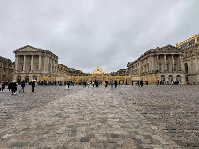 Exterior view of the Palace of Versailles featuring its ornate golden gates, adorned with intricate detailing and royal symbols, gleaming in the sunlight against the grand palace façade.