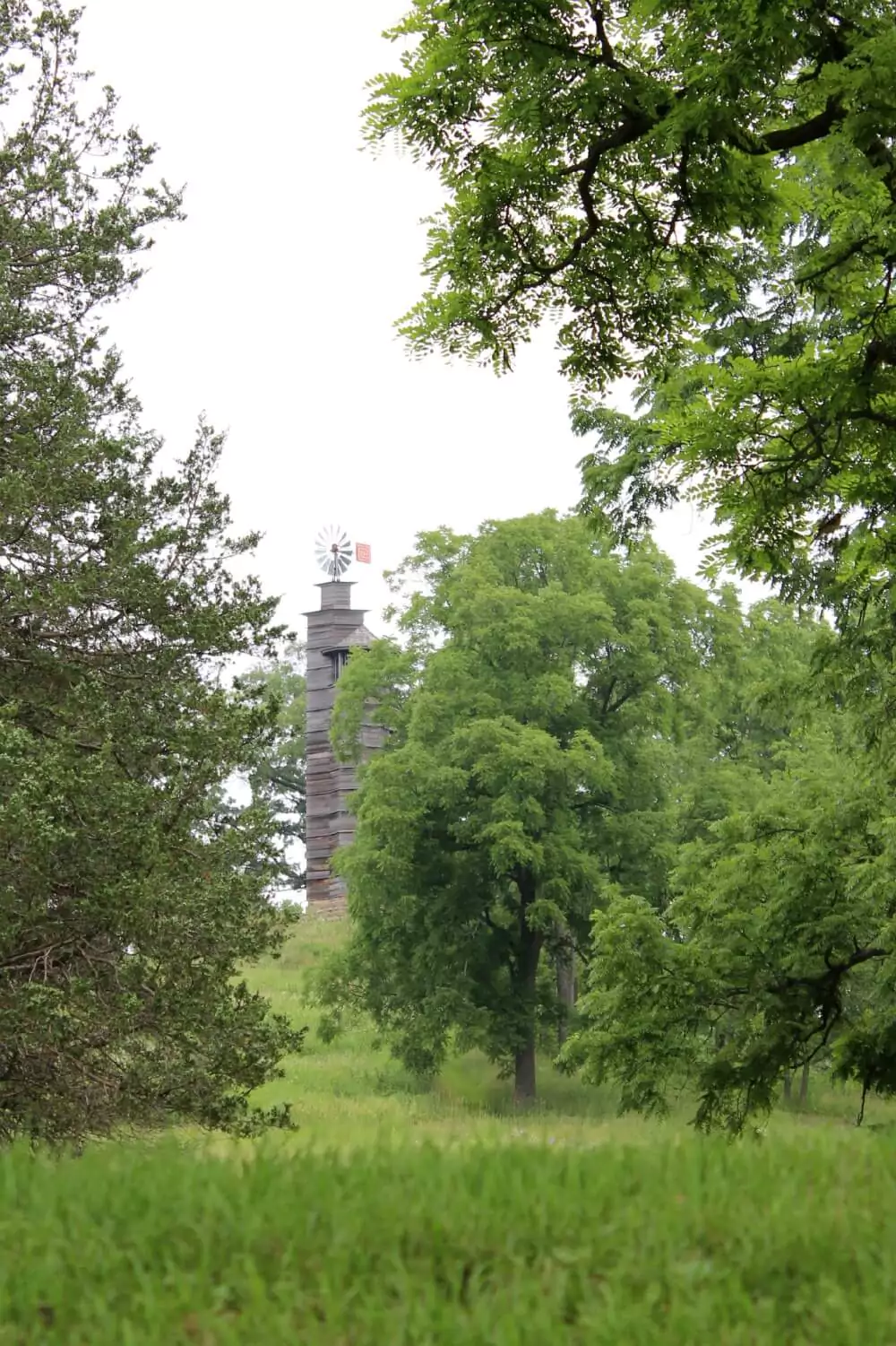 A tall, weathered wooden windmill tower rises above a grassy hill, partially hidden by lush green trees. The windmill’s metal blades and red tail stand out against the overcast white sky.