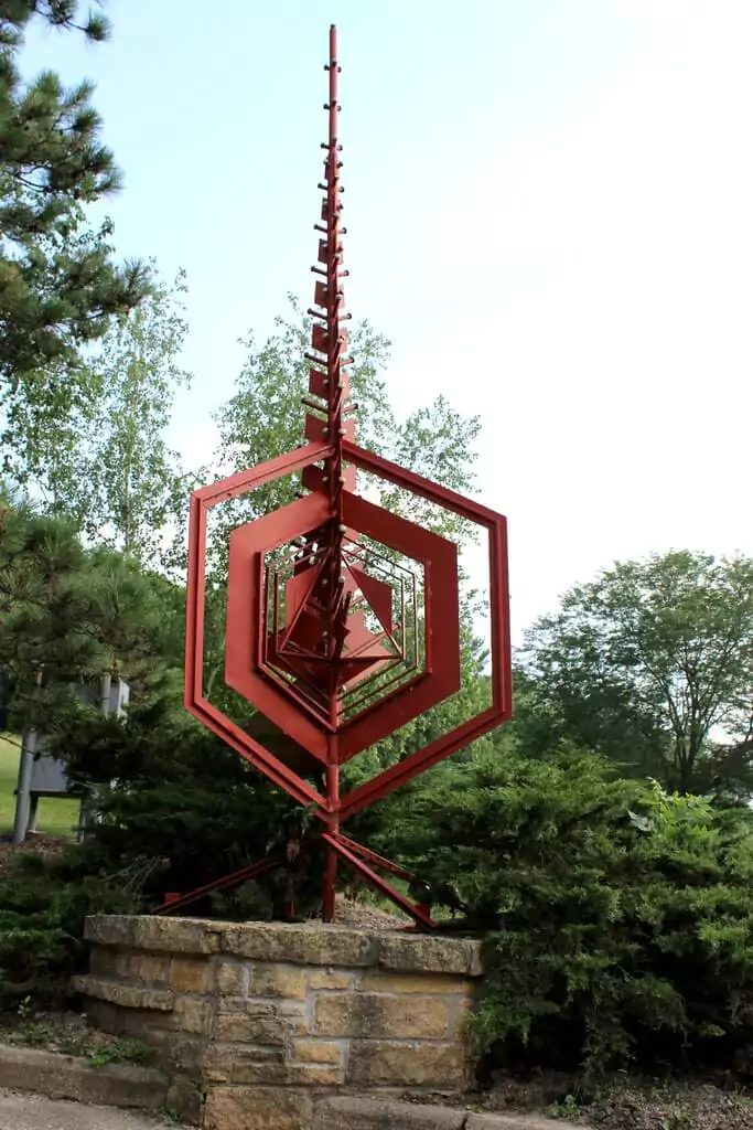 A tall red metal sculpture with layered geometric hexagon shapes narrowing to a pointed tip, mounted on a stone base and surrounded by green shrubs and trees. The angular design creates a sense of upward motion against the pale blue sky.