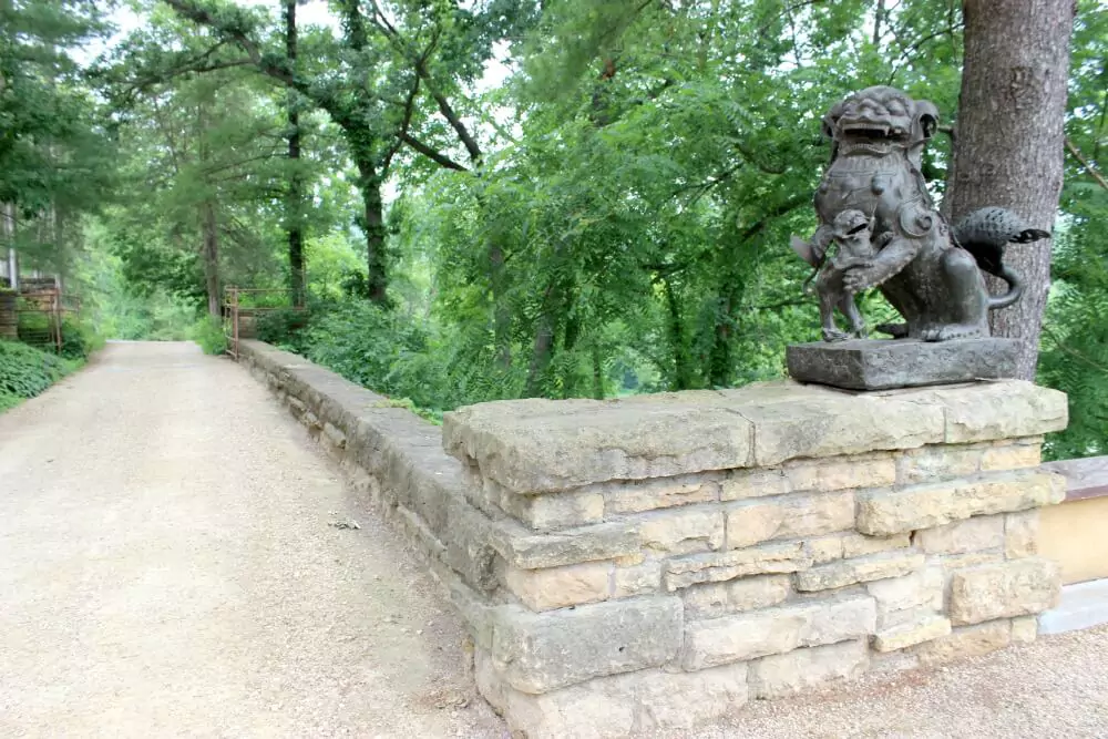 A stone wall lines a gravel pathway through a wooded area, with a sculpted guardian lion statue perched at the corner. The dark, intricately detailed figure faces outward, framed by dense green foliage in the background.