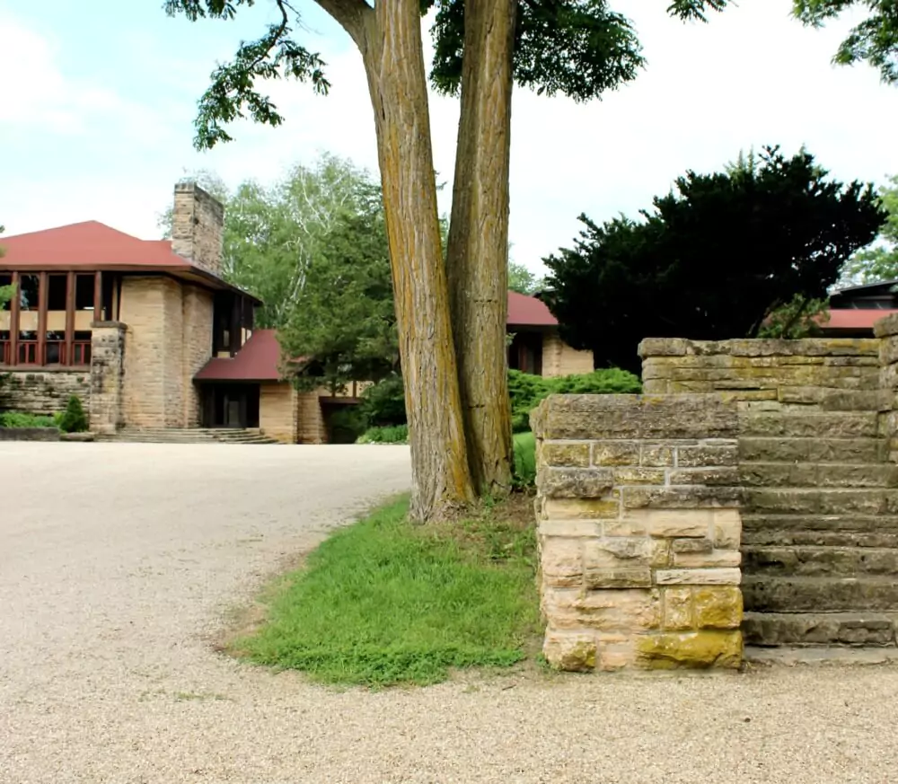 Stone steps bordered by a short stone wall lead up beside two tall trees, with a large tan brick building featuring red roofs and wide windows in the background. The scene is framed by green grass, shrubs, and a bright, partly cloudy sky.