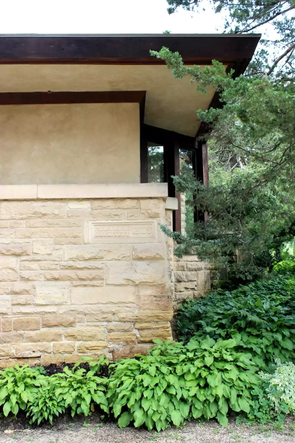 Close-up of a tan stone and stucco building corner with a carved inscription on the wall, partially shaded by overhanging evergreen branches. Lush green plants grow along the base, contrasting with the structure’s earthy tones.