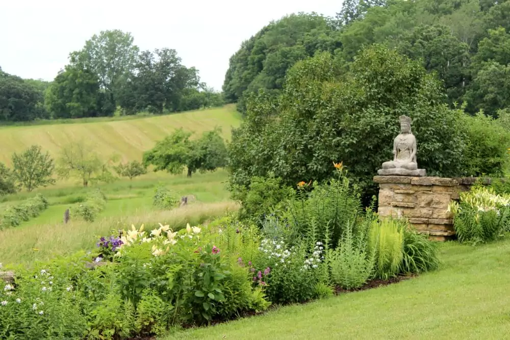 A stone statue of a seated figure rests atop a short stone wall, overlooking a lush garden with blooming flowers in shades of white, yellow, pink, and purple. Beyond the garden, rolling green fields and scattered trees stretch toward a wooded hillside.