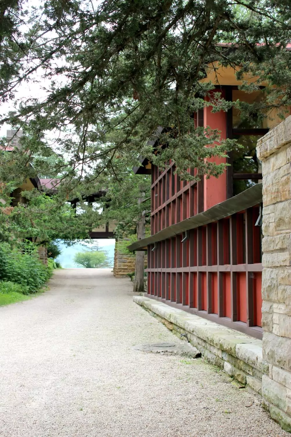 A gravel pathway passes between tan stone walls and a tall red-and-brown grid-patterned structure, with tree branches arching overhead. In the distance, the path leads to an open view framed by an overhead walkway and lush greenery.