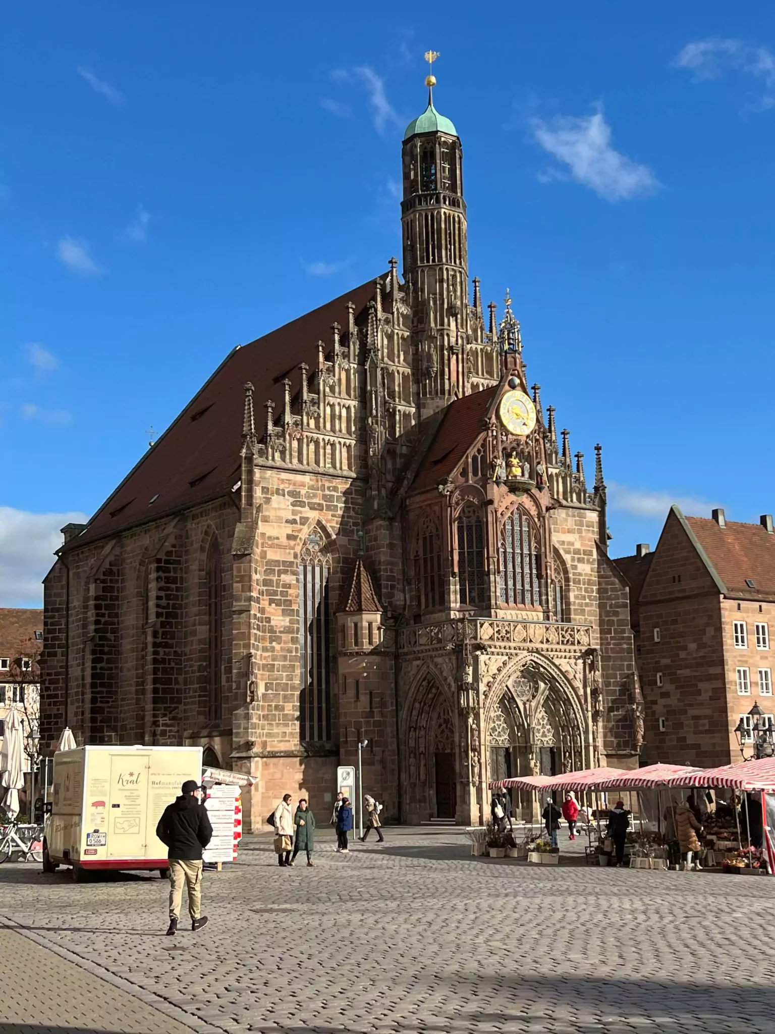 The Frauenkirche overlooks the Hauptmarkt in Nuremberg, Germany, where market vendors line the square with striped red-and-white stalls. The Gothic church’s intricate facade towers over the lively scene, blending history with the everyday bustle of local trade.