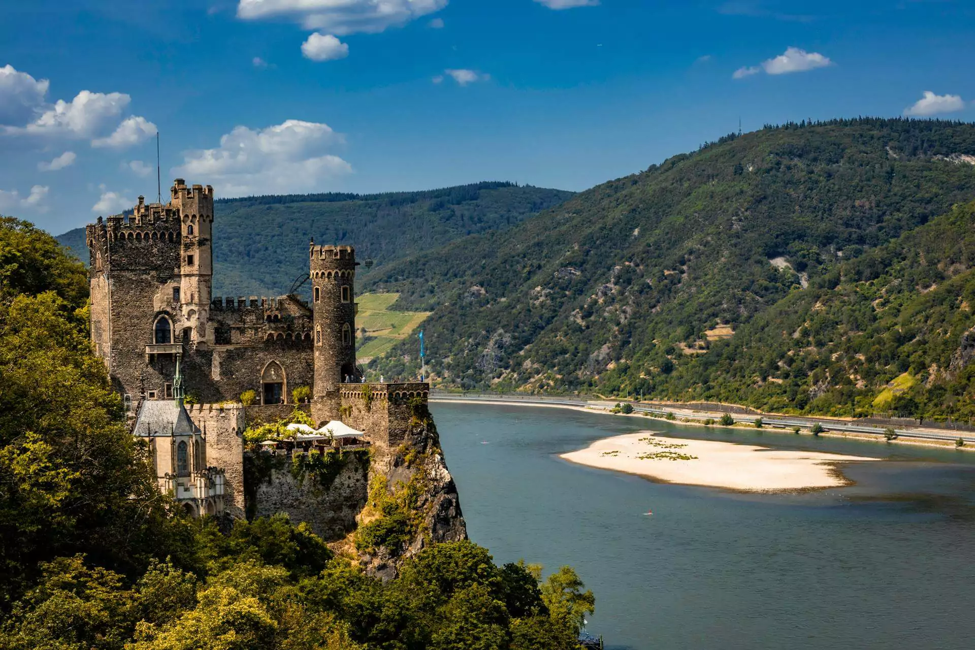 Burg Rheinstein in Germany, a small but dramatic castle clinging to a rocky cliff above the Rhine River, with Romanesque-style architecture, turrets, and a lush garden courtyard.