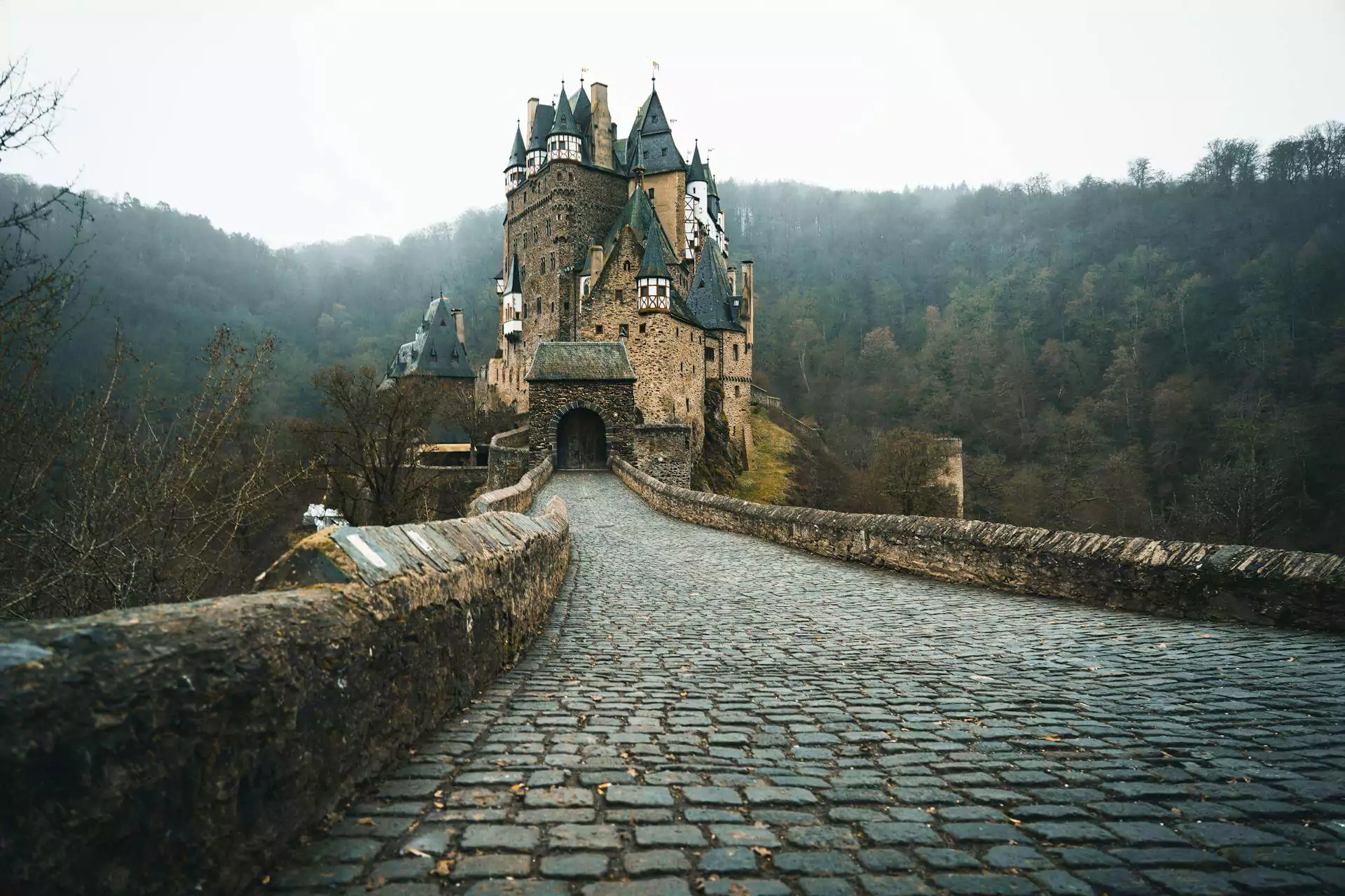 Eltz Castle in Germany, a well-preserved medieval fortress nestled in a wooded valley, featuring spired towers, half-timbered upper levels, and stone walls rising above the Elzbach River.