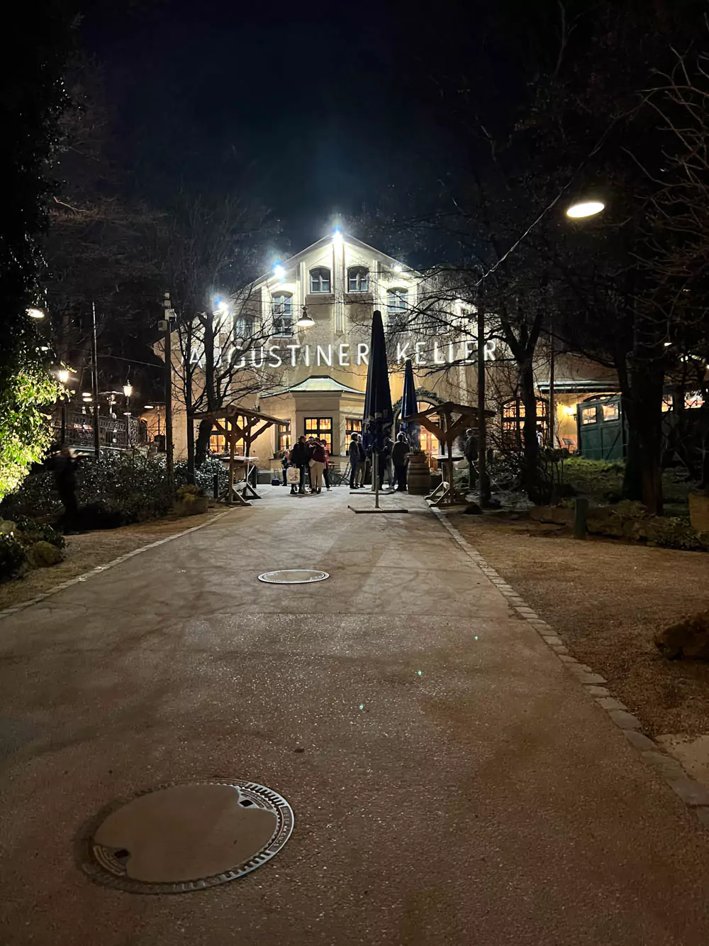 The lively exterior of Augustiner-Keller beer garden in Munich illuminated with warm lights at night, with patrons gathered around outdoor tables under festive string lighting. The historic building’s traditional Bavarian architecture creates a cozy, inviting atmosphere.