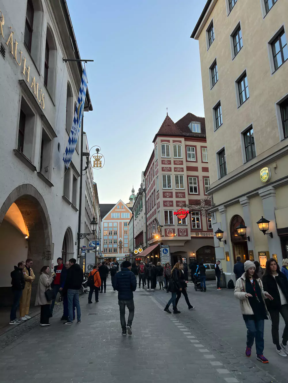 A crowded street scene outside Munich’s Hofbräuhaus beer hall with pedestrians, tourists, and outdoor seating under umbrellas. The historic building’s traditional Bavarian facade anchors the lively urban atmosphere.