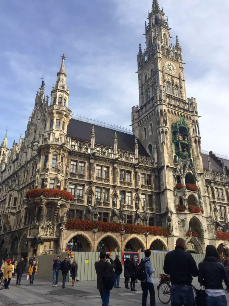 The New Town Hall (Neues Rathaus) at Marienplatz in Munich, Germany, featuring ornate Gothic Revival architecture with arched windows, statues, and the iconic Glockenspiel tower rising above the bustling square.